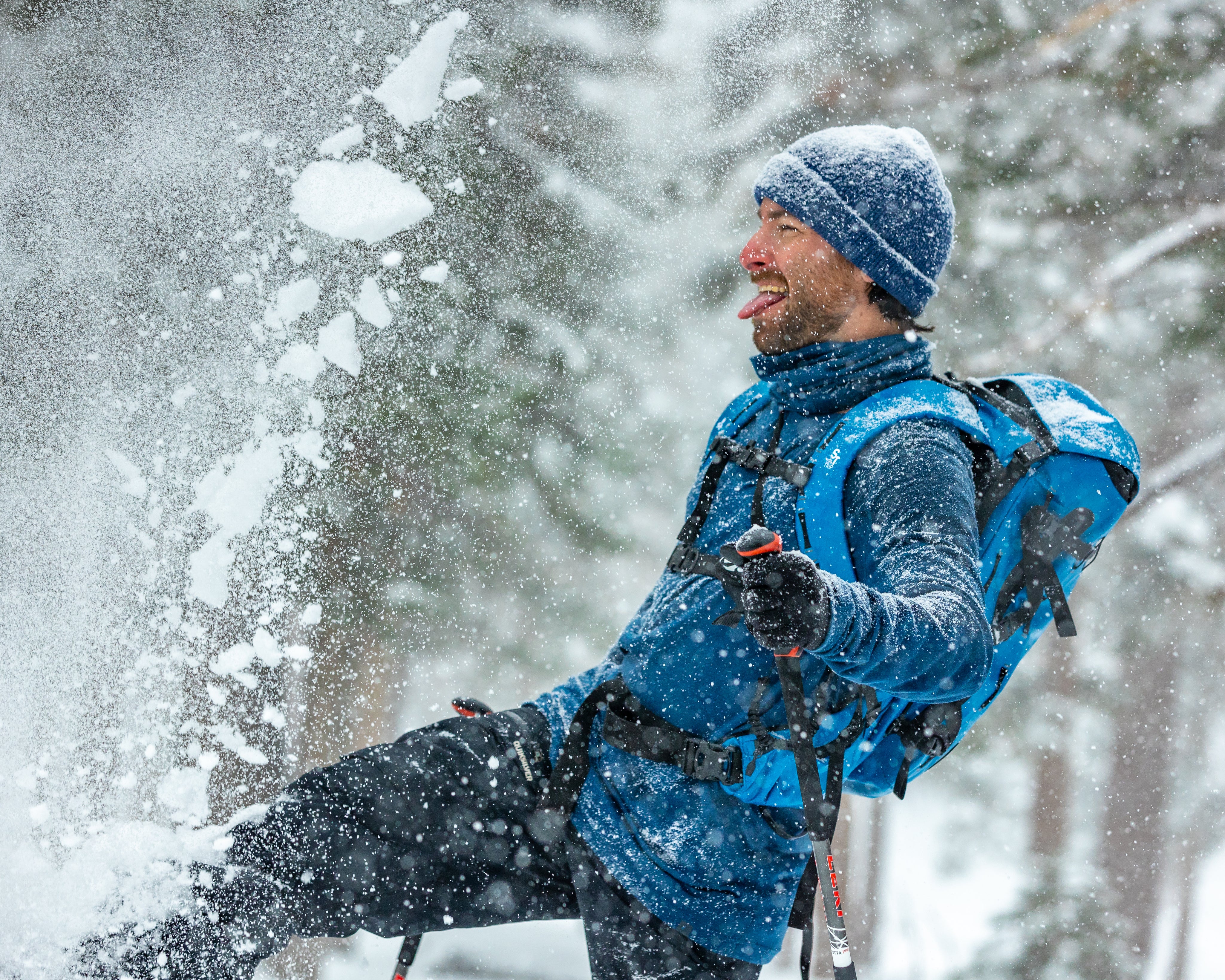 a man smiles kicking up snow while wearing blue Ridge Merino layers and a blue backpack