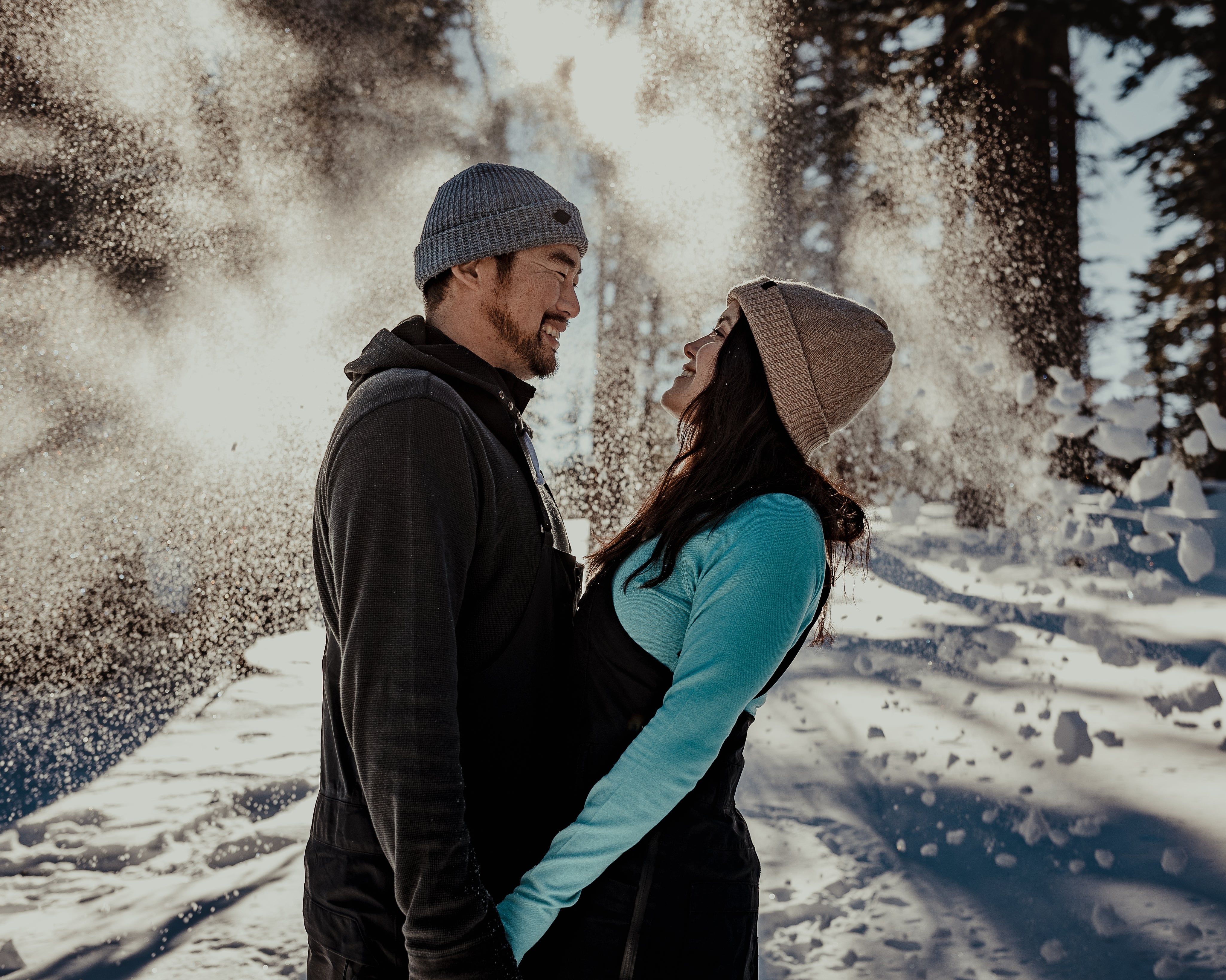 a couple wearing Ridge Merino winter layers smiles at each other while sun pours through the snowy trees in the background