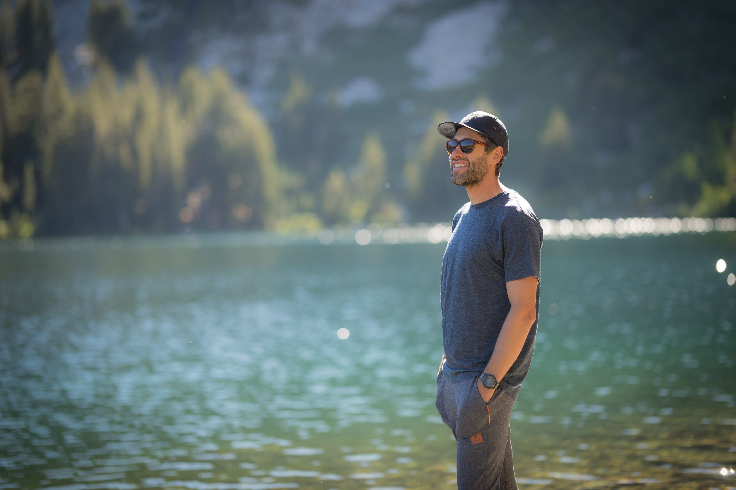 A man smiles beside an alpine lake wearing a Ridge Natural Tencel Tee and sunglasses