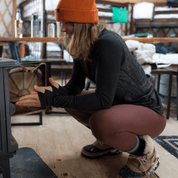 a woman warms up by a wood stove in a backcountry yurt wearing Ridge base layers
