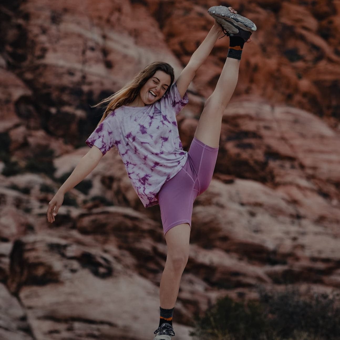a woman sticks her tongue out doing a standing split wearing Hilltop Shorts and a tie-dye Wander Tee
