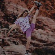 a woman sticks her tongue out doing a standing split wearing Hilltop Shorts and a tie-dye Wander Tee