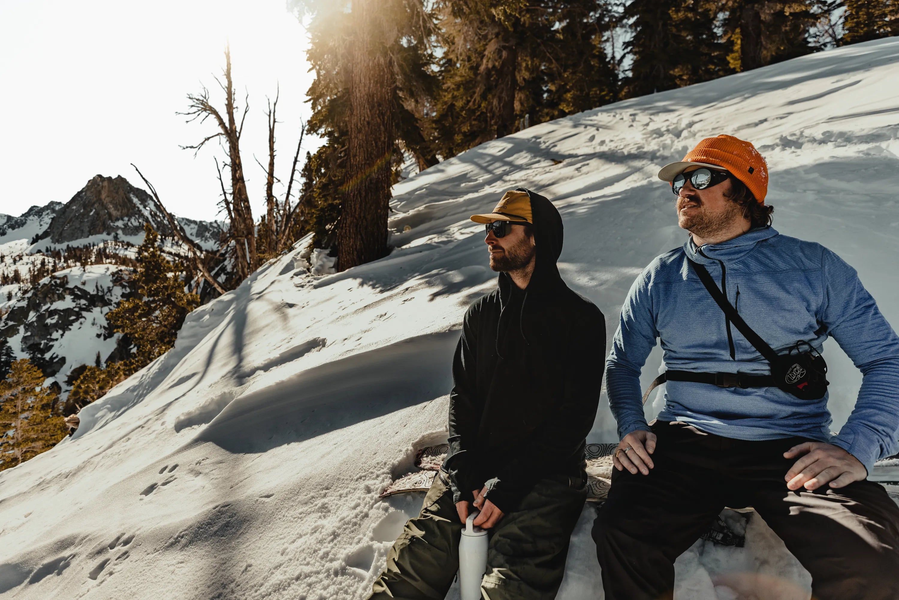 two men wearing Ridge Merino hoodies sit on a snowboard while taking a break on a splitboarding tour