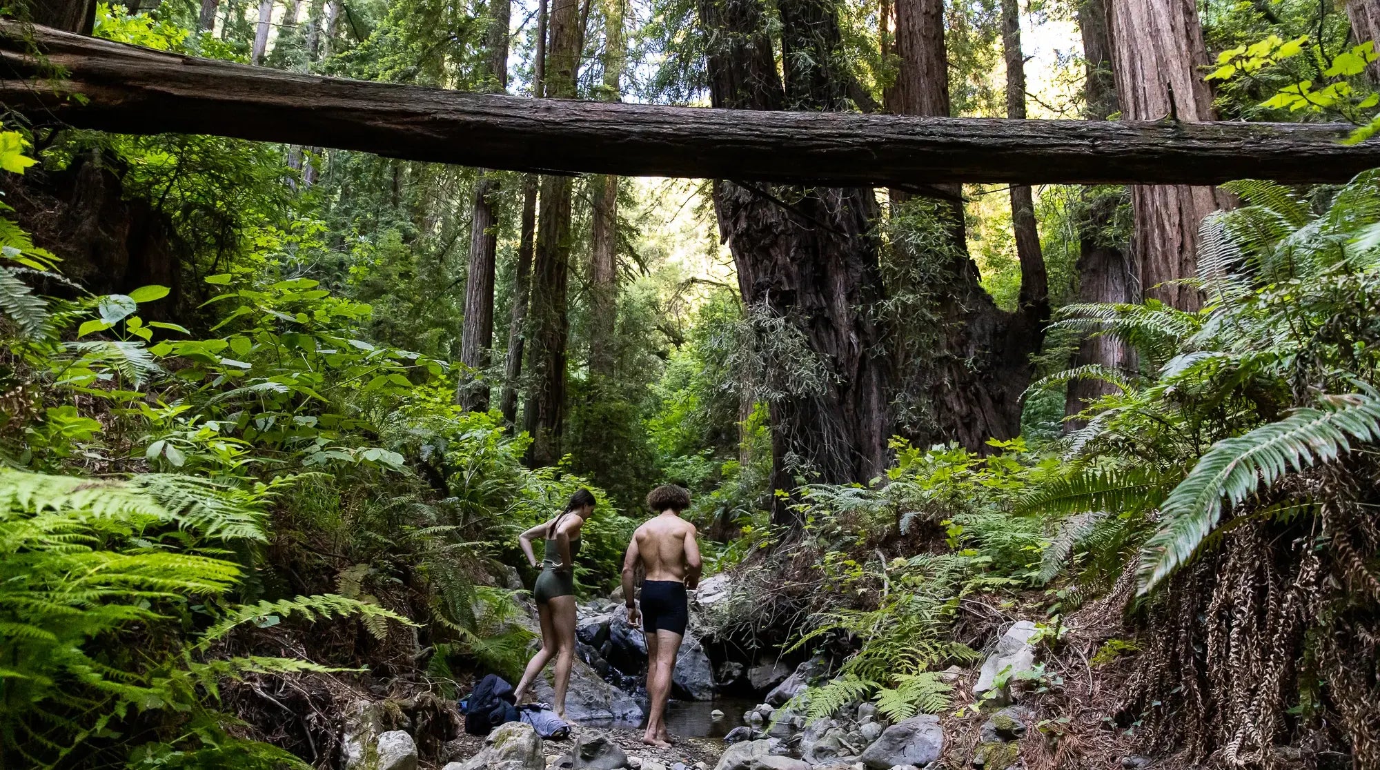 a man and a woman walk through the woods to a swimming hole in their Ridge Merino underwear