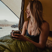 A woman smiles in a tent wearing the Merino wool bralette and drinking coffee