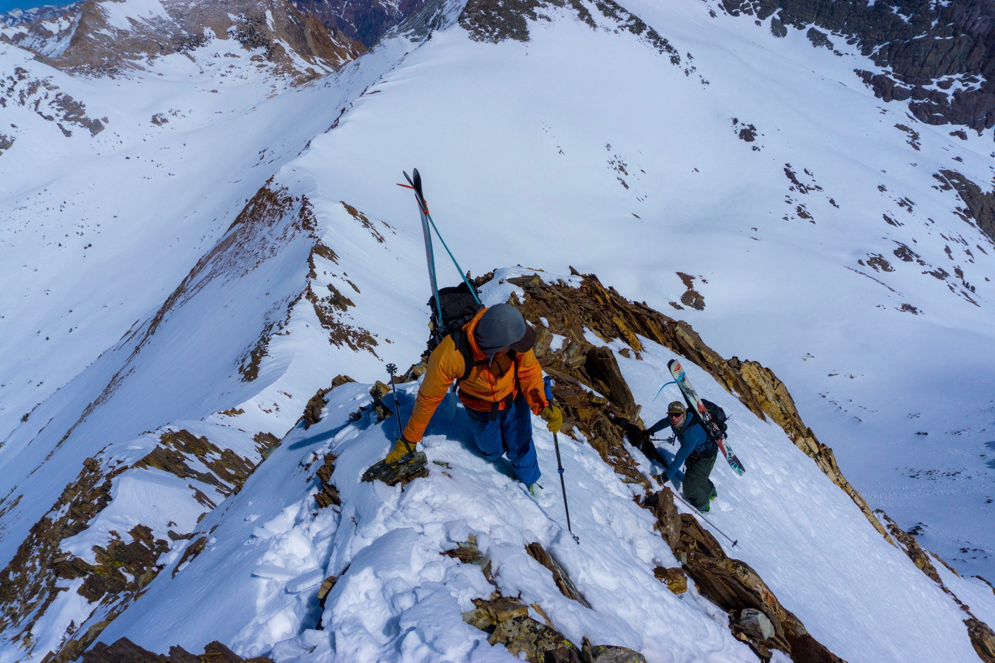 two people climbing up a mountain in the snow with skis on their backs