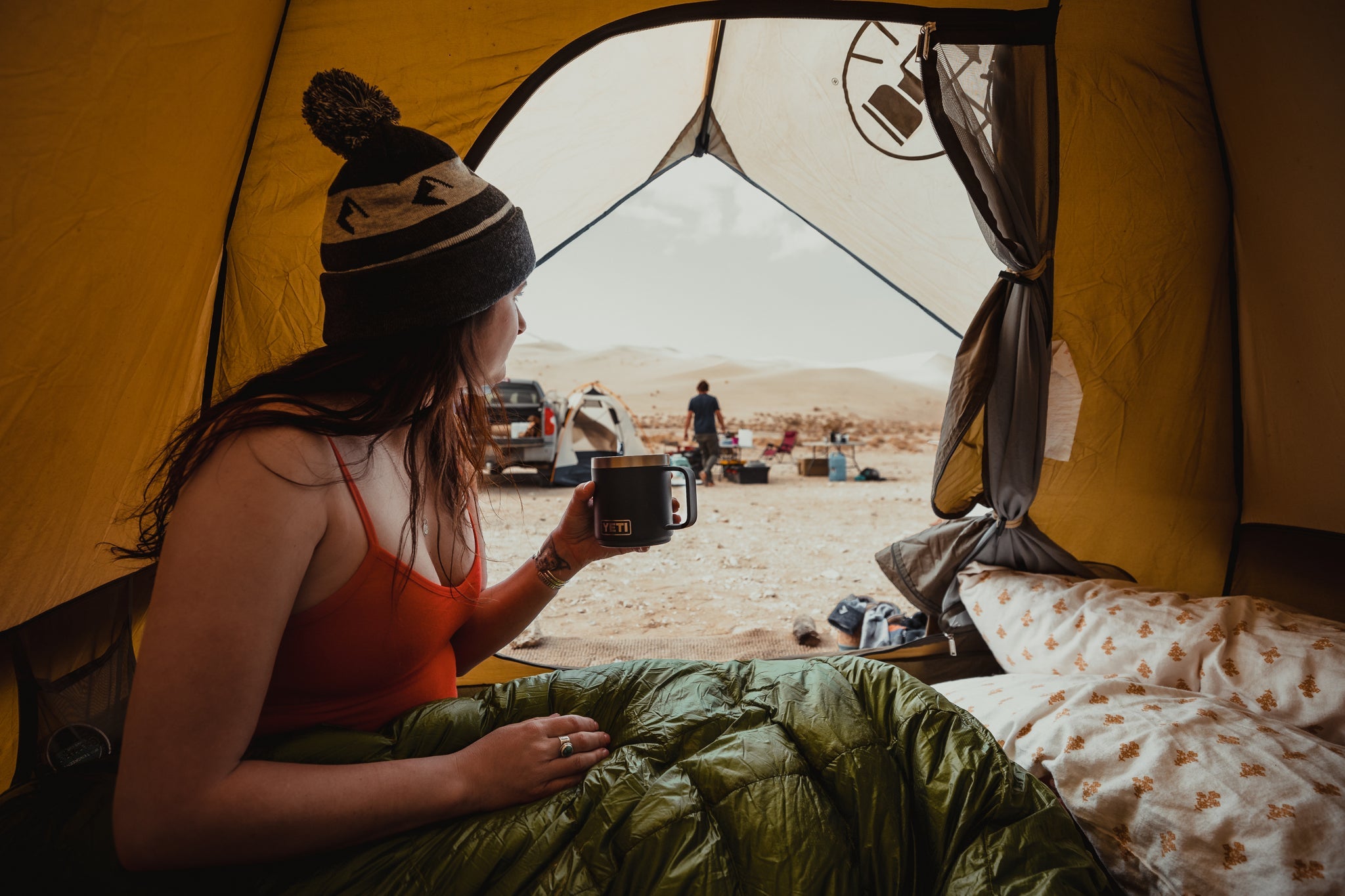 a woman drinking morning coffee in a tent wearing a red Ridge Merino Bralette and Ridge Beanie