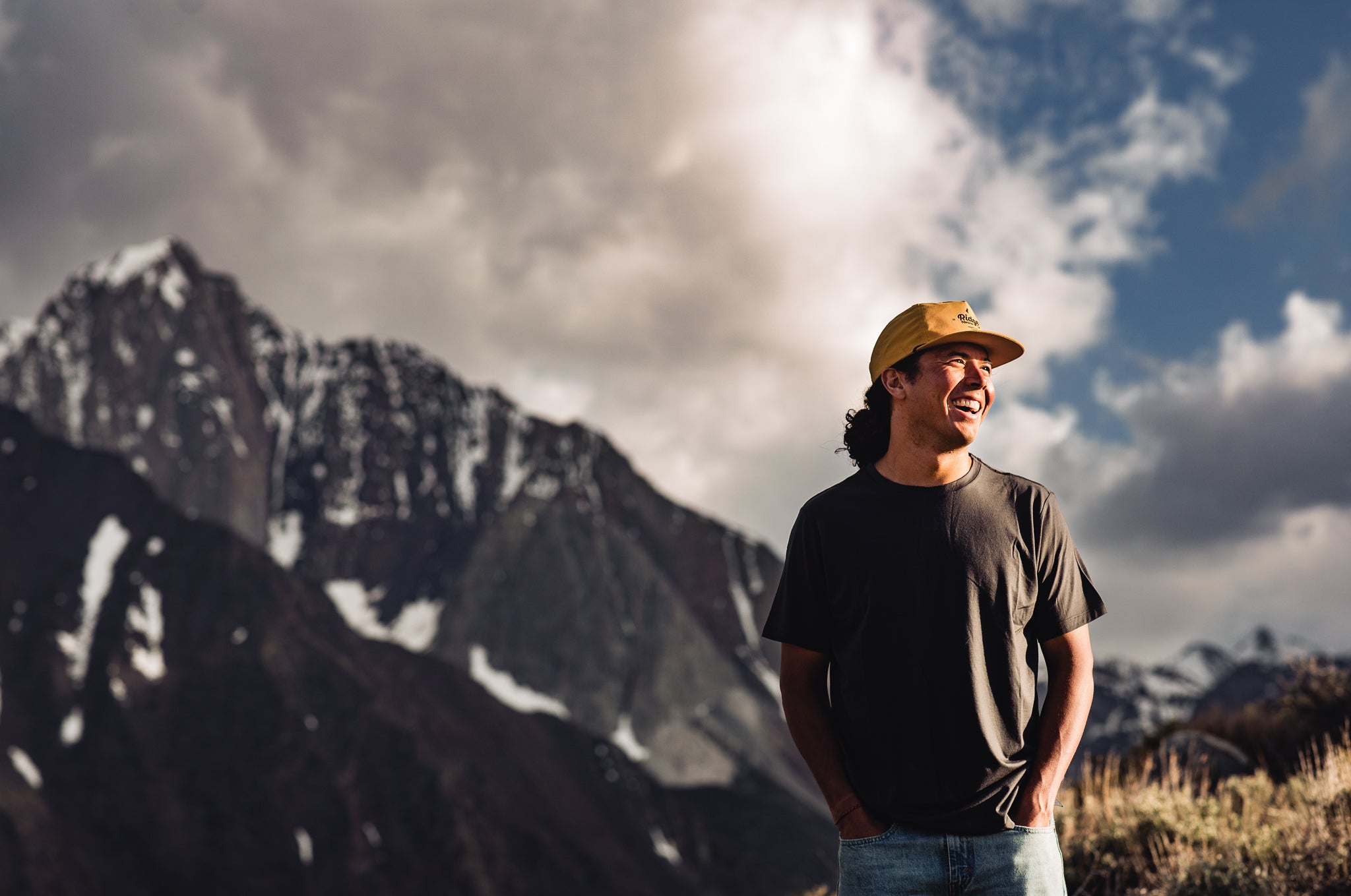 a man laughs in front of Mt. Morrison wearing a Journey Merino T-shirt and a yellow Ridge Merino hat