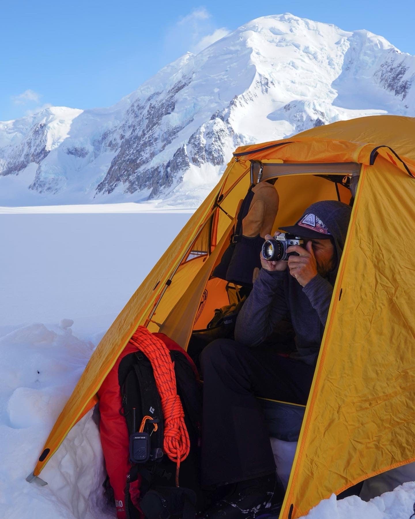 Nick Cahill photographs a scene from a yellow tent set up in snowy mountains