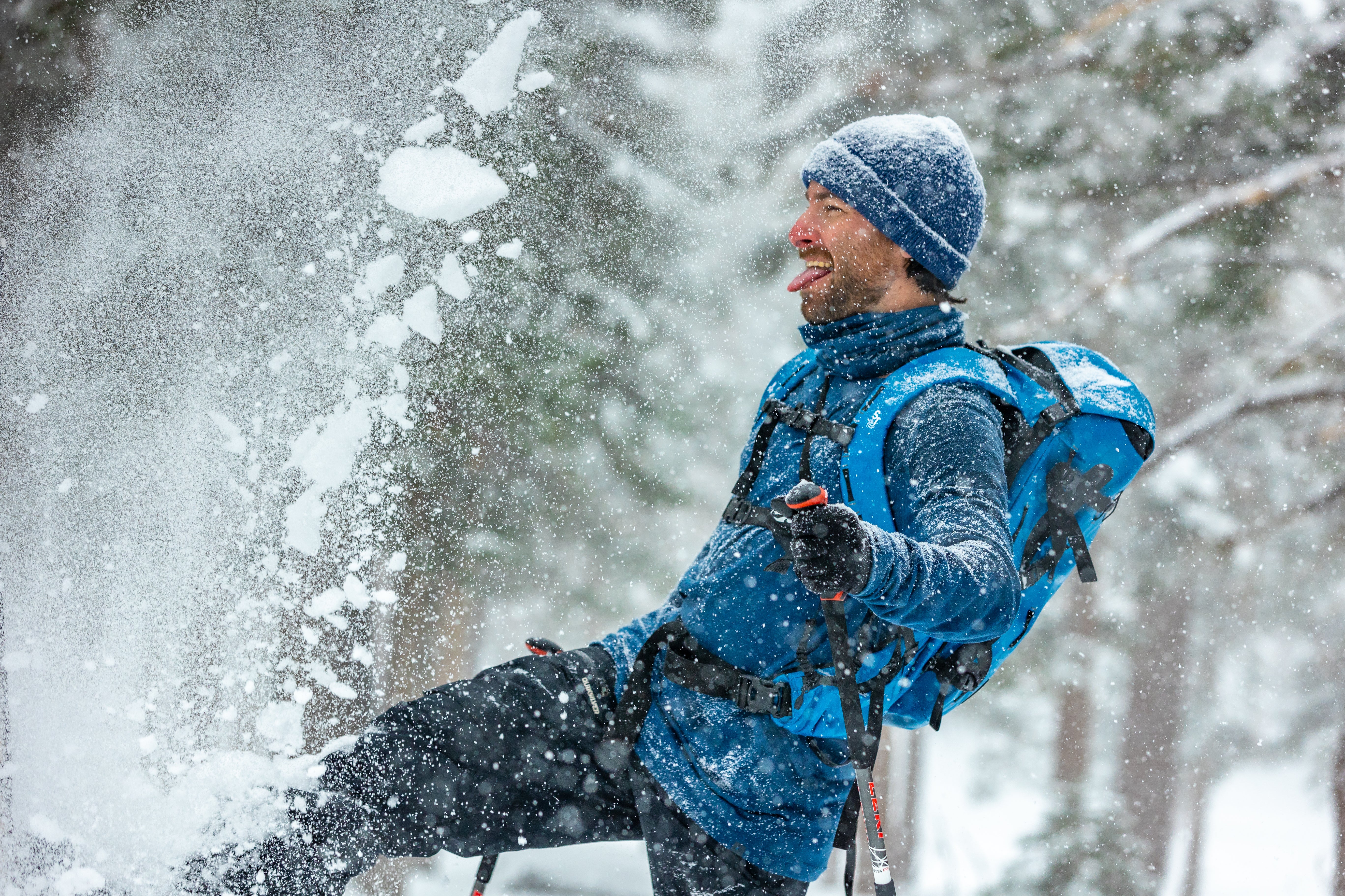 a man smiles kicking up snow while wearing blue Ridge Merino layers and a blue backpack