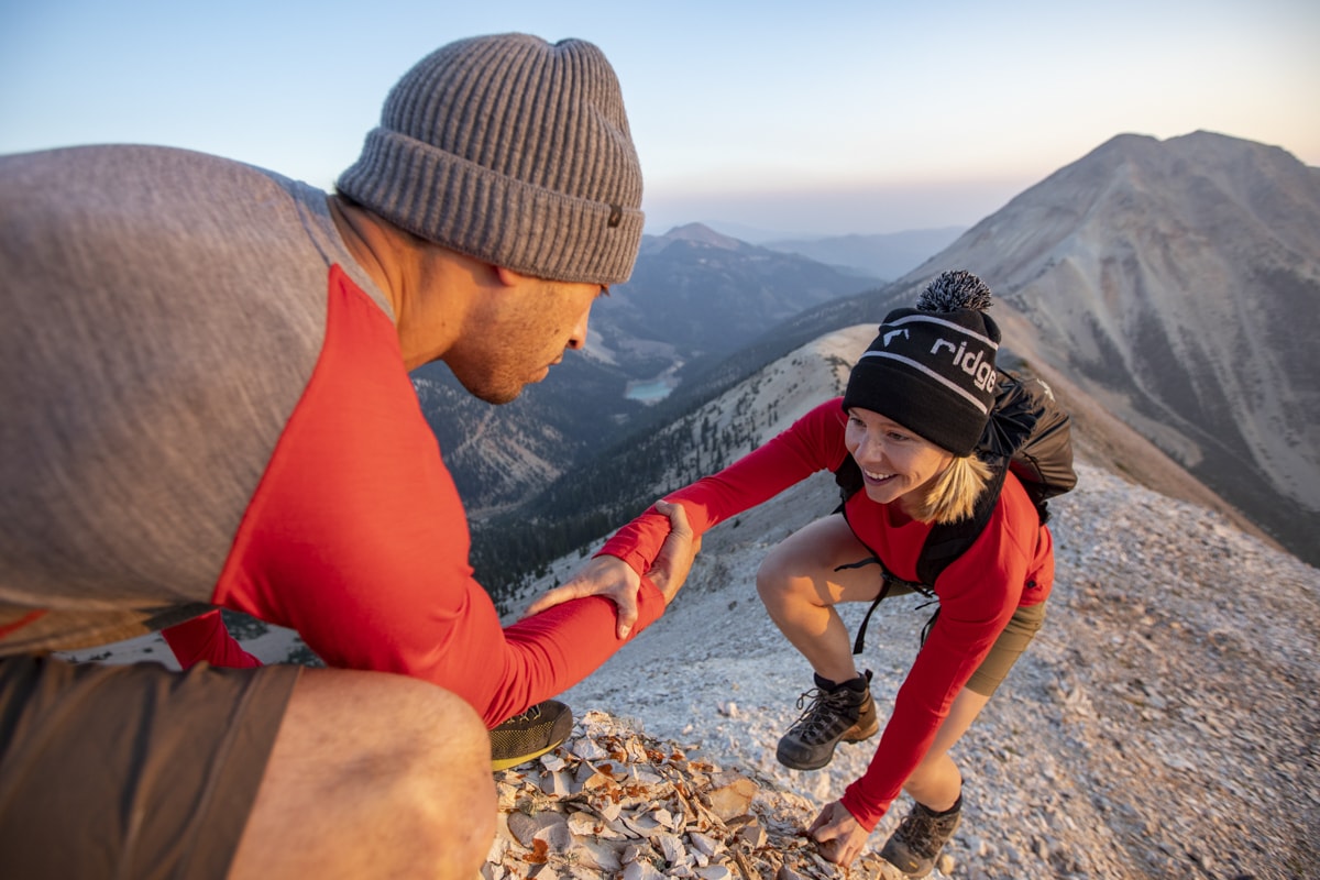 one hiker helps another get to the summit, both wearing red Ridge Aspect base layers