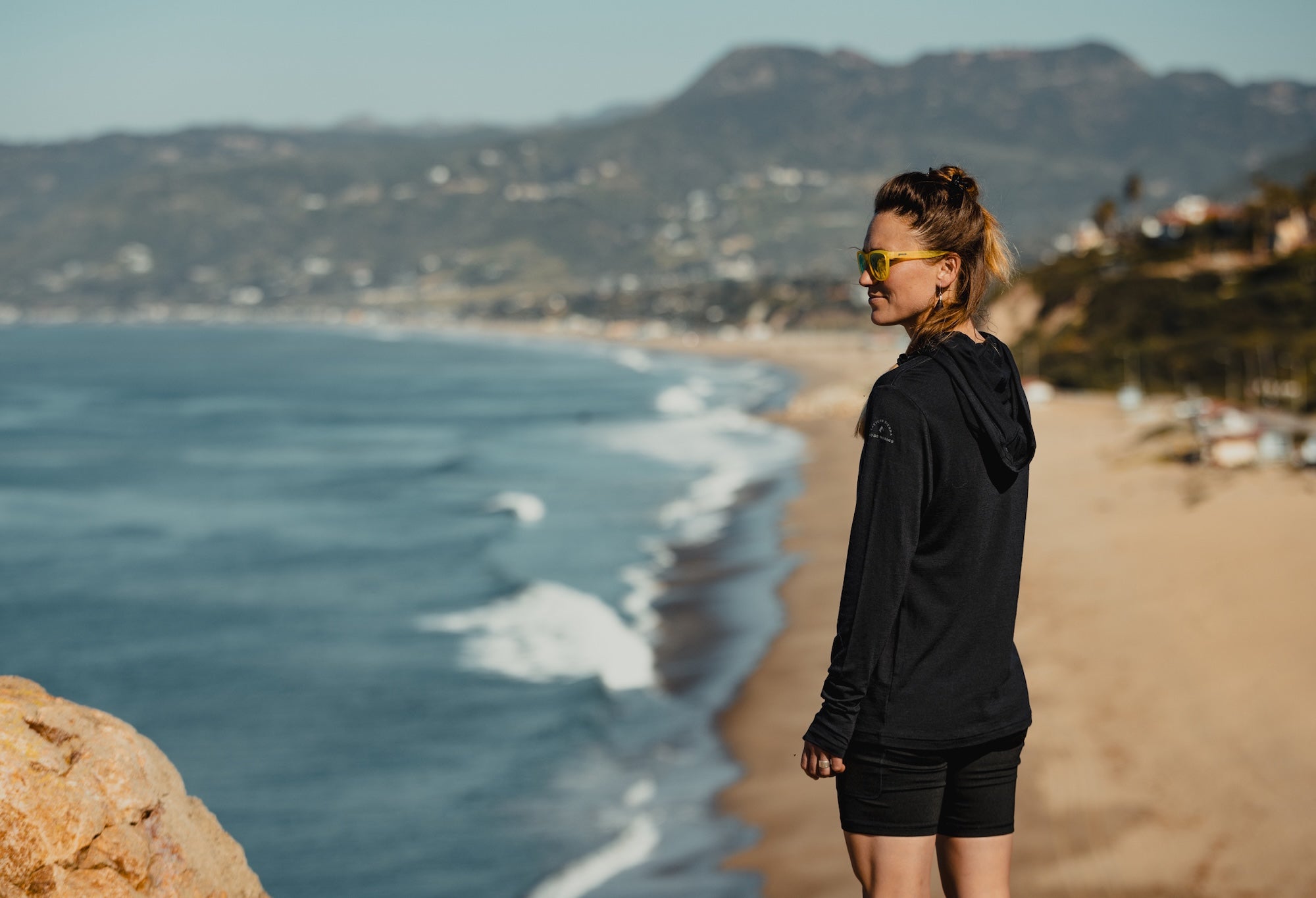 a woman smiles standing above a beach wearing a black Solstice Sun Hoodie and black Hilltop Shorts and sunglasses