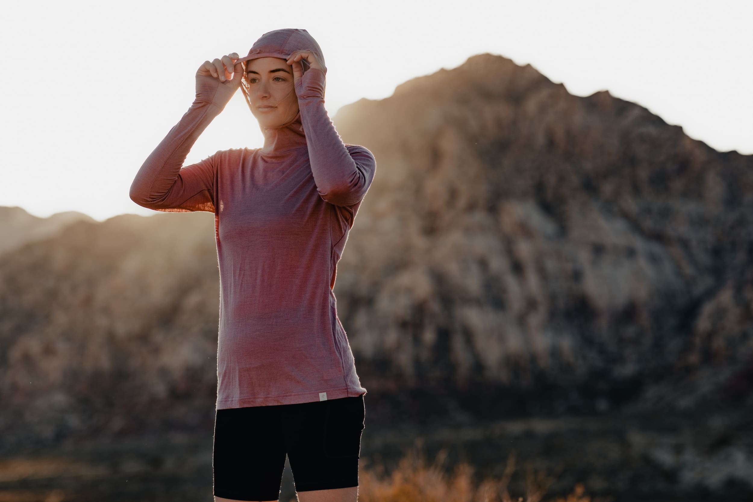a woman pulling the hood up on a pink Pursuit Ultralight Hoodie as the sun sets behind her