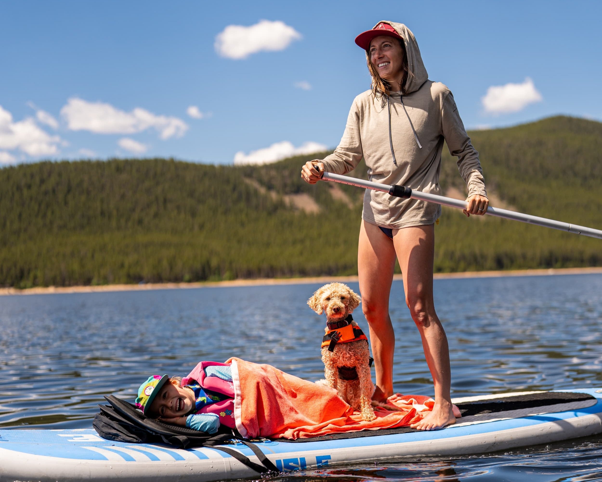 a mom smiles stand up paddleboarding wearing a Solstice Sun Hoodie while her child and dog sit on the paddleboard