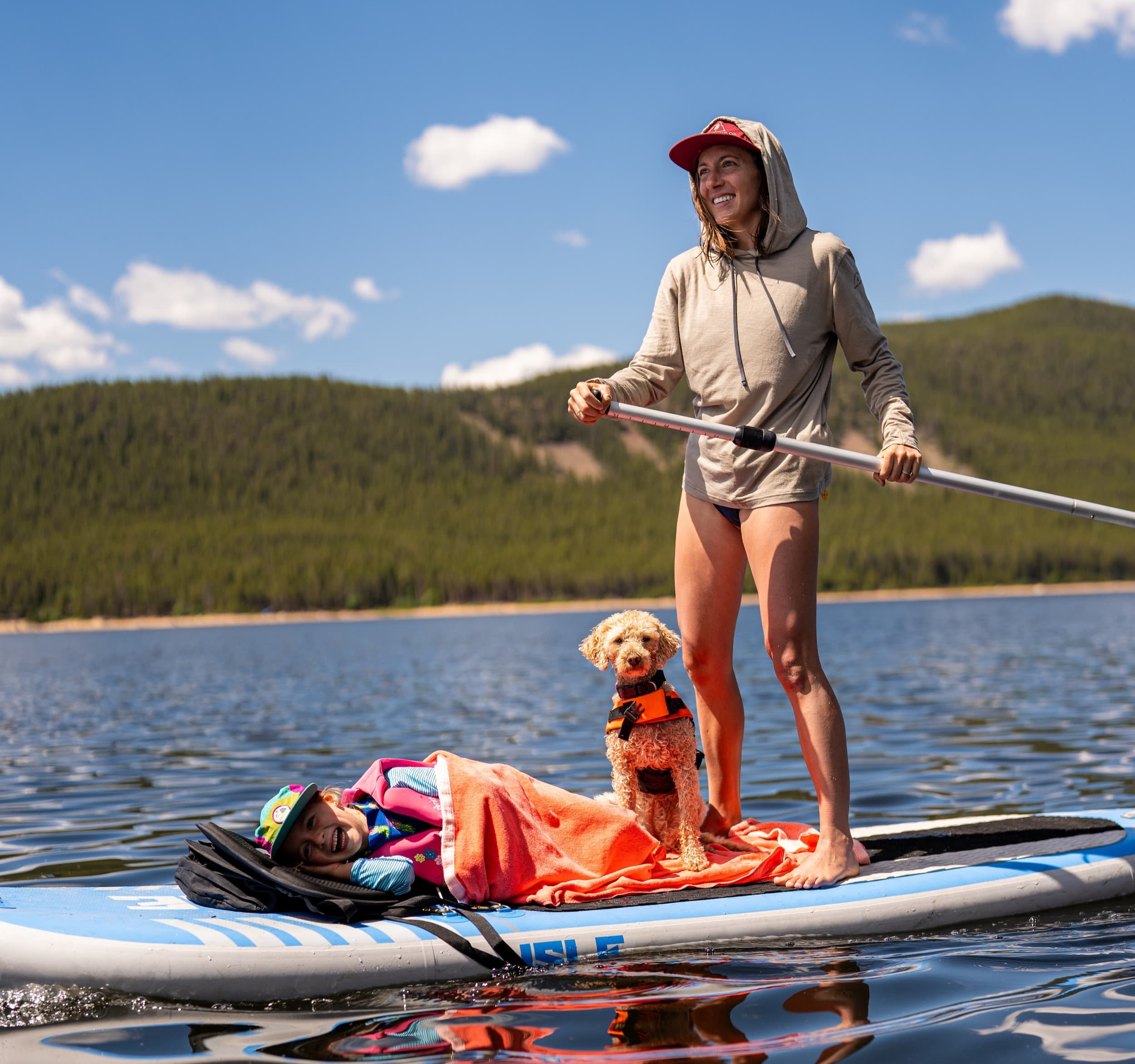 a mom smiles stand up paddleboarding wearing a Solstice Sun Hoodie while her child and dog sit on the paddleboard