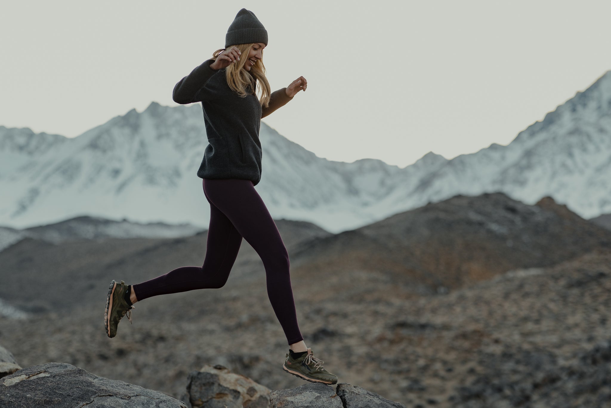 a woman hops between rocks in the Eastern Sierra wearing Ridge Merino layers with snowy mountains in the background