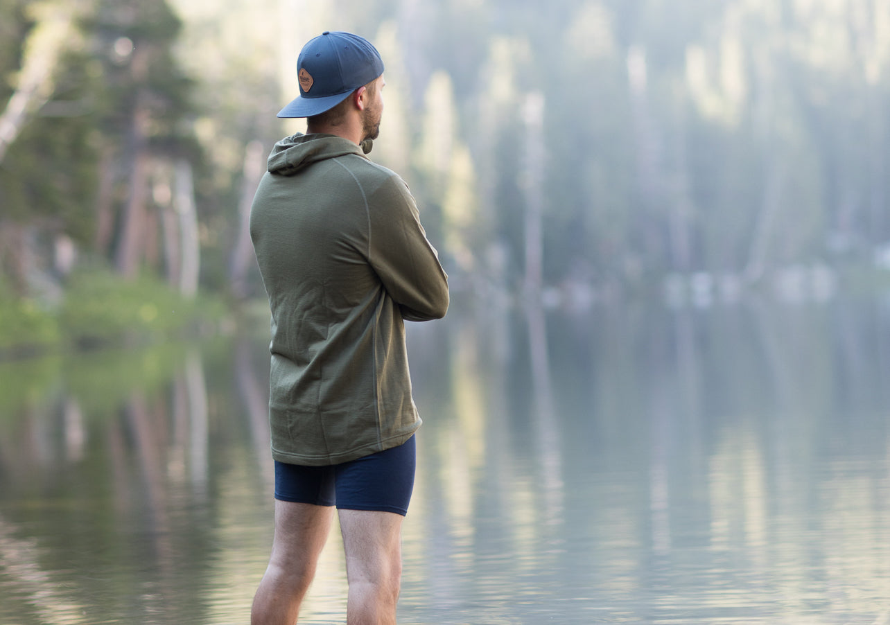a man standing by an alpine lake in a Ridge Merino hoodie and Ridge Boxer Briefs