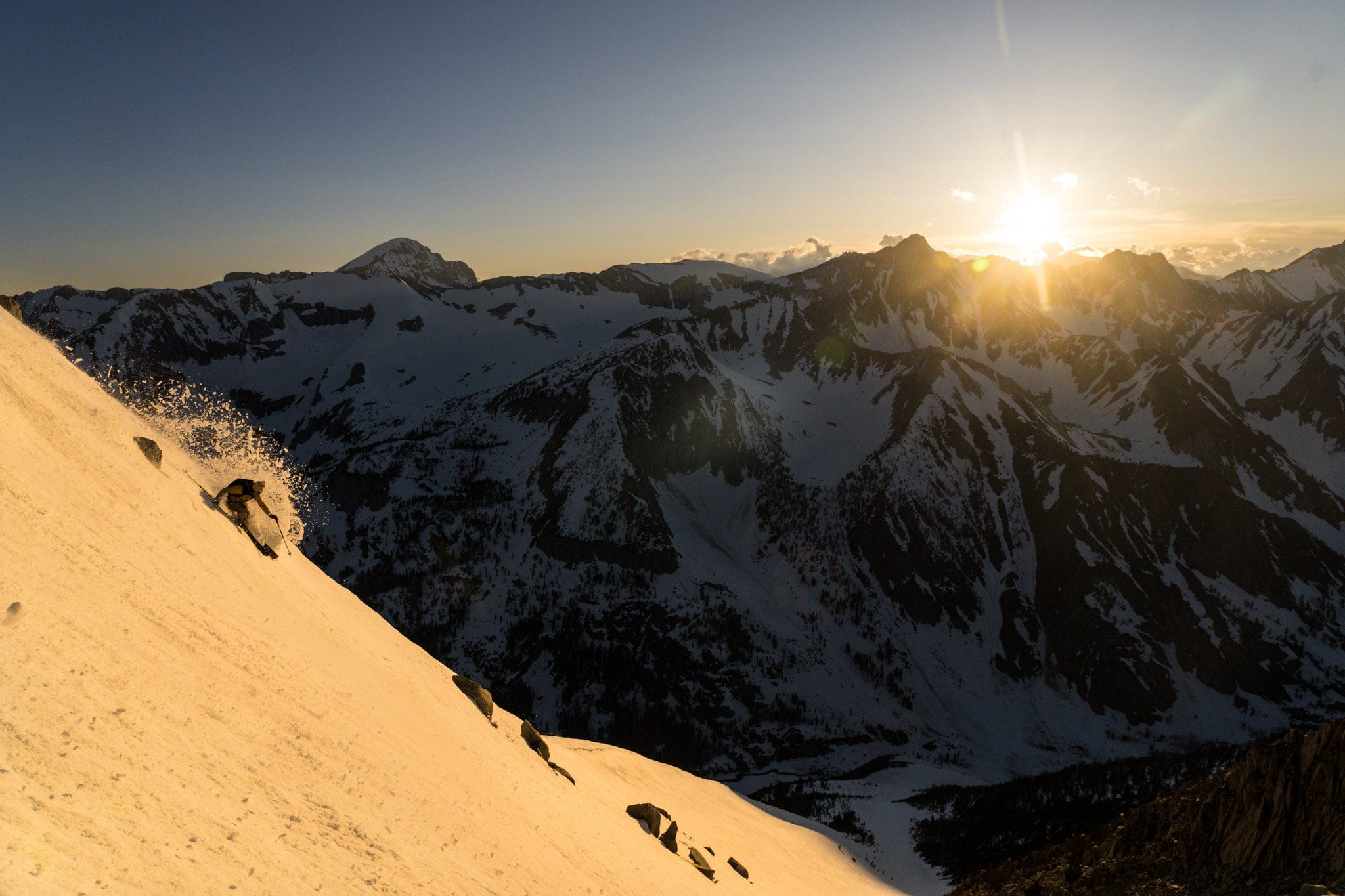 A skier skis down a ski run in Mammoth Lakes while the sun sets over mountains the background and casts golden light on the ski run