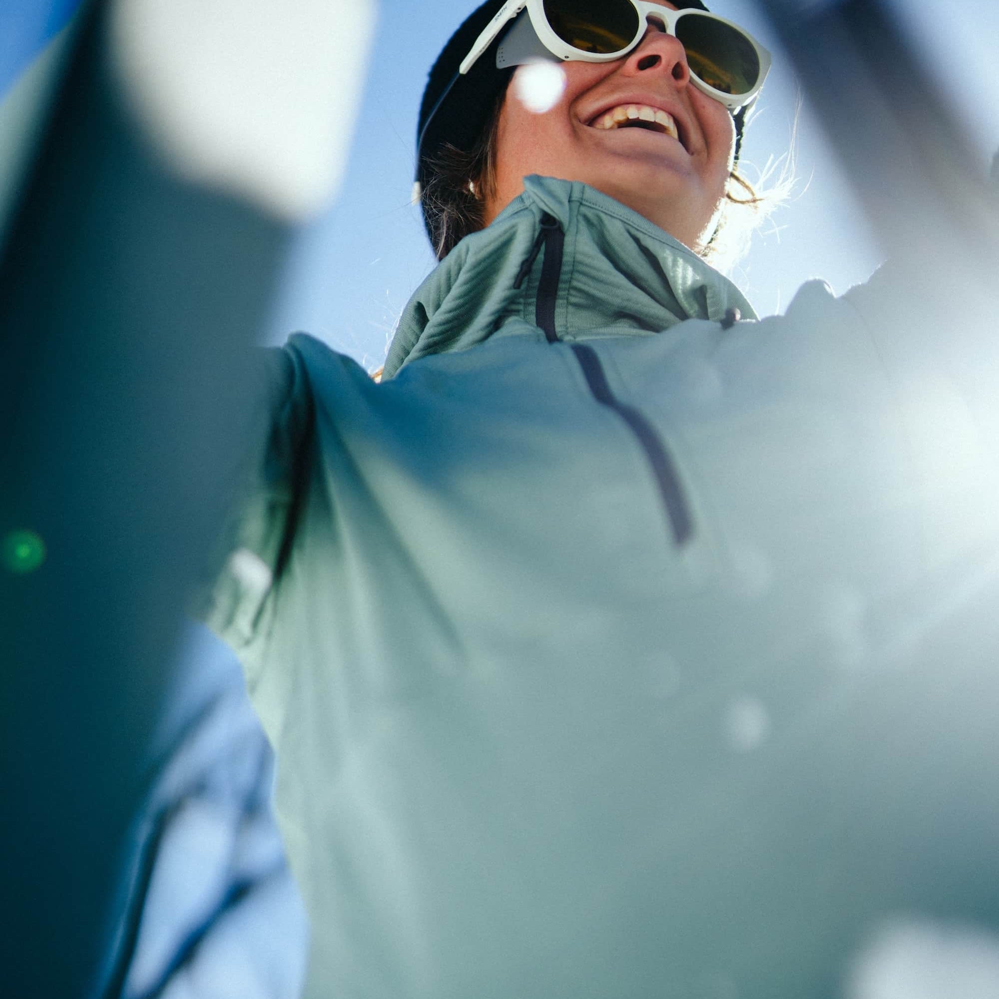 a woman smiling adjusting her skis wearing an Iceberg Green Convict Canyon Hoodie and white sunglasses