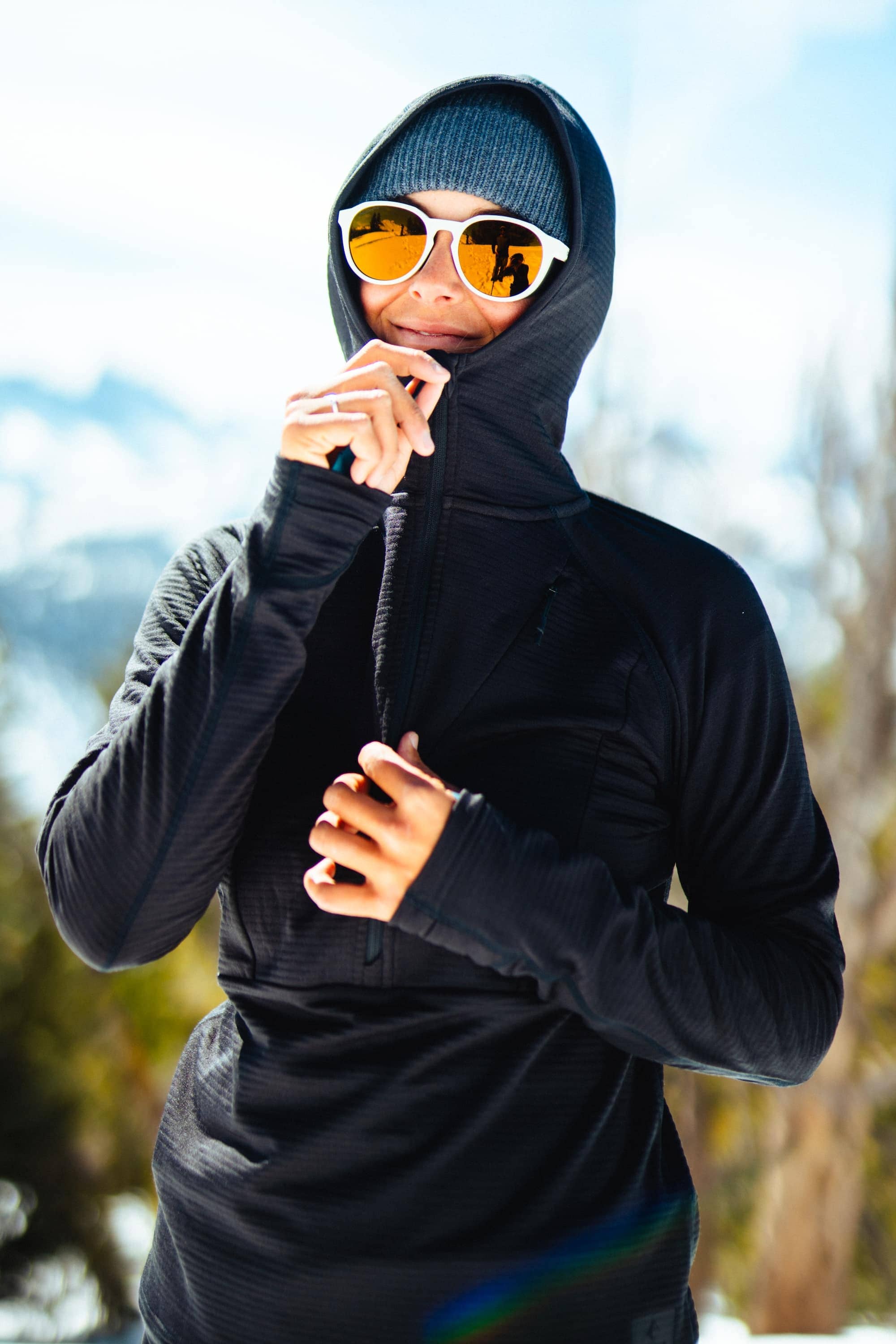 a woman wearing sunglasses smiling zipping up a Convict Canyon Hoodie in black on a sunny winter day