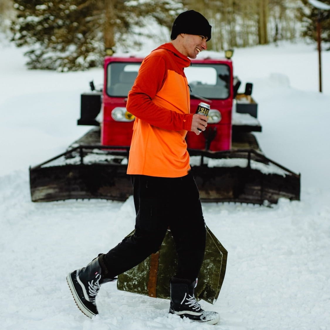 a man carries firewood in front of a snow cat wearing black Cloudripper Joggers and an orange Convict Canyon Hoodie