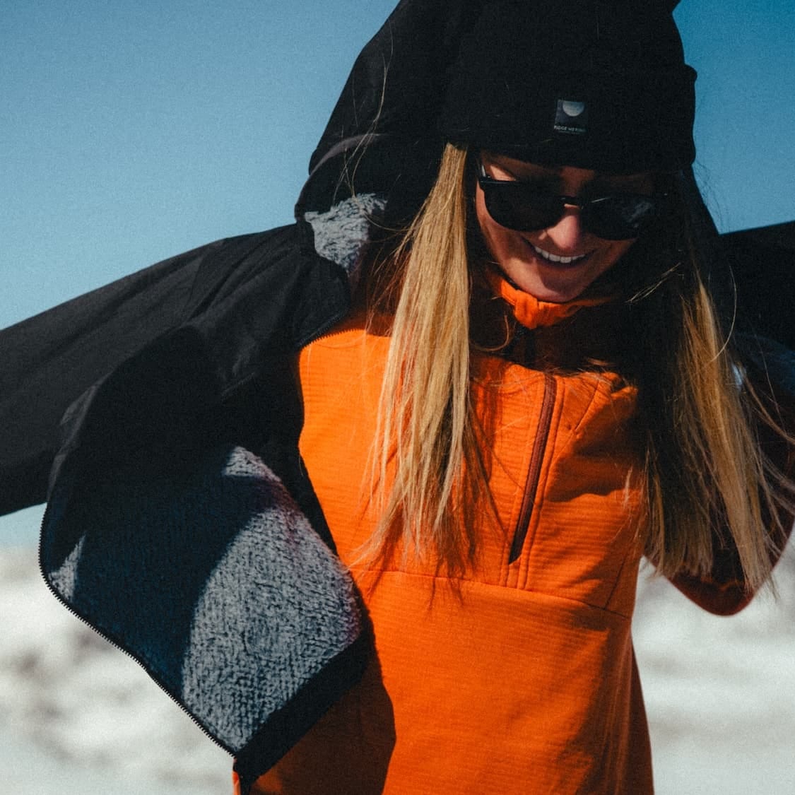 a woman putting on an Alpha Insulated Wool Jacket over an orange Convict Canyon Hoodie