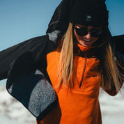 a woman putting on an Alpha Insulated Wool Jacket over an orange Convict Canyon Hoodie