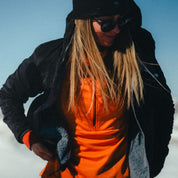 a woman putting on an Alpha Insulated Wool Jacket over an orange Convict Canyon Hoodie
