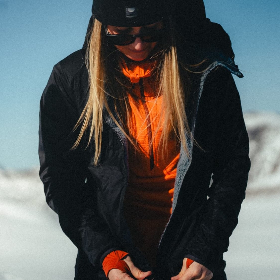 a woman putting on an Alpha Insulated Wool Jacket over an orange Convict Canyon Hoodie