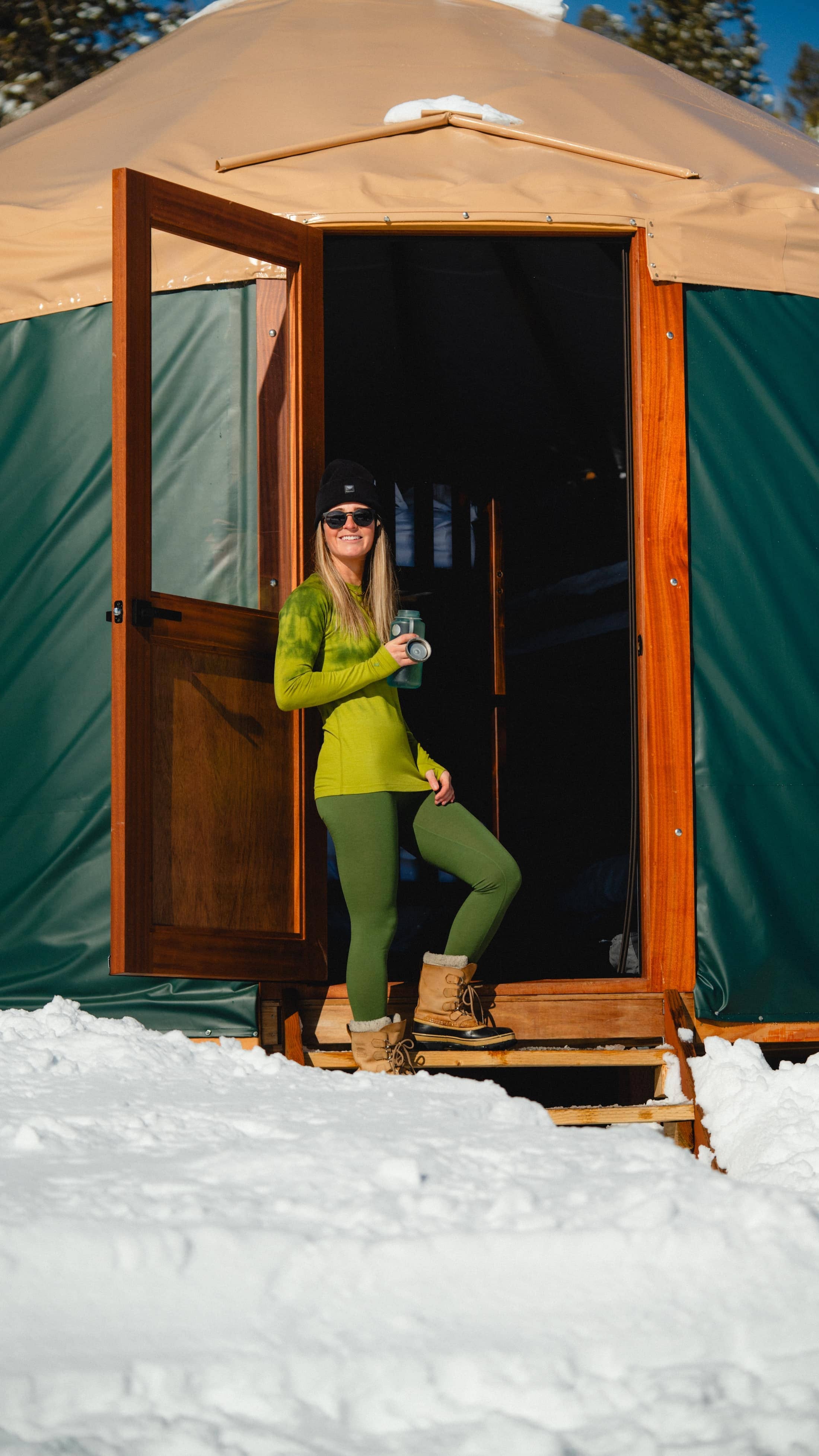 a woman smiles in front of a backcountry ski yurt wearing Aspect Midweight Merino Base Layers