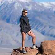 a woman in a black windbreaker standing on rocks on a mountain hike