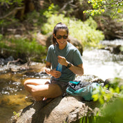 a woman taking a break from hiking by a stream in a Journey Merino Wool T-Shirt