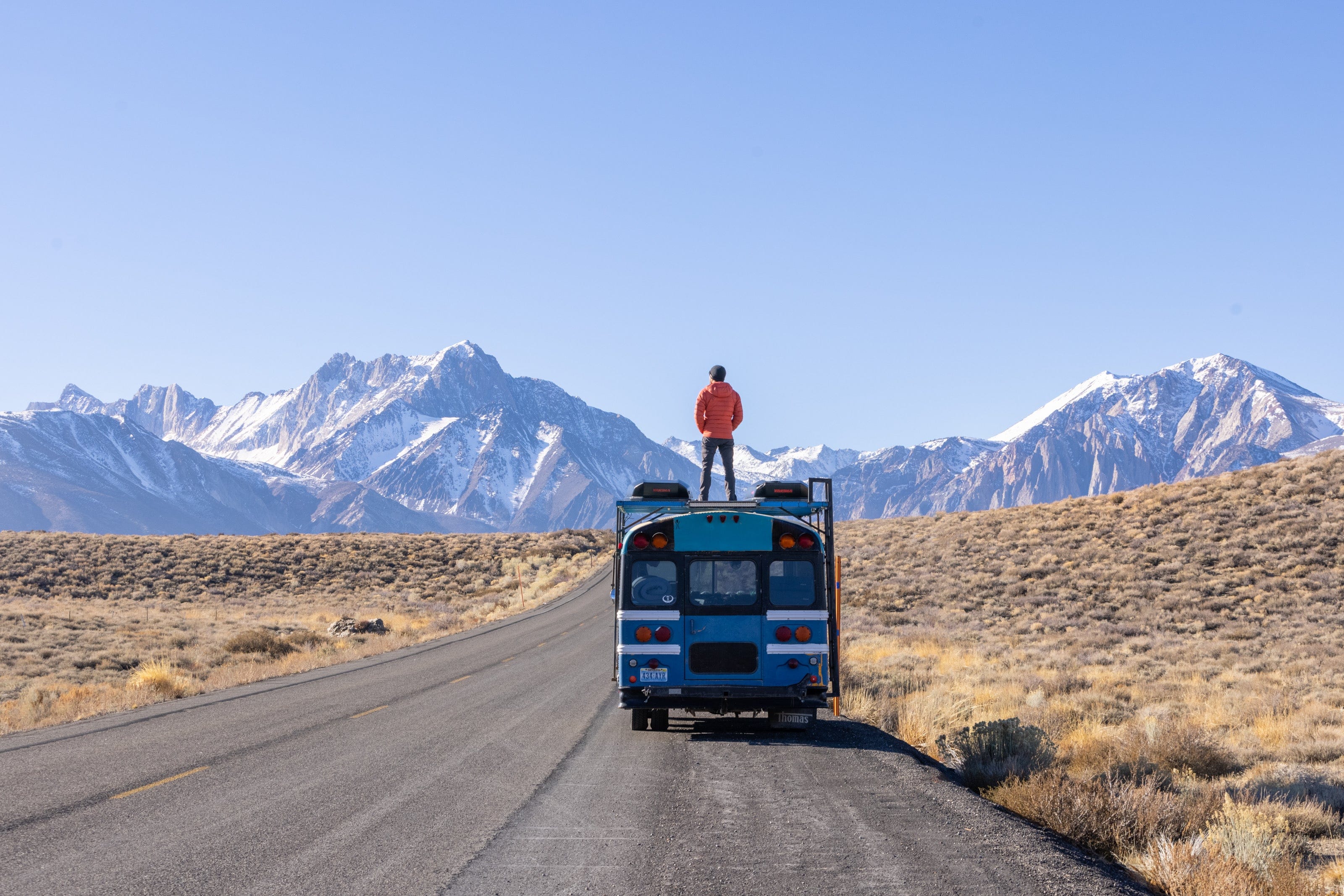 a man stands on top of his converted school bus he lives in with sweeping snowy mountains in Mammoth Lakes in the background