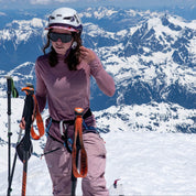 a woman adjusting her ski helmet in the backcountry wearing a pink ultralight Pursuit Hoodie