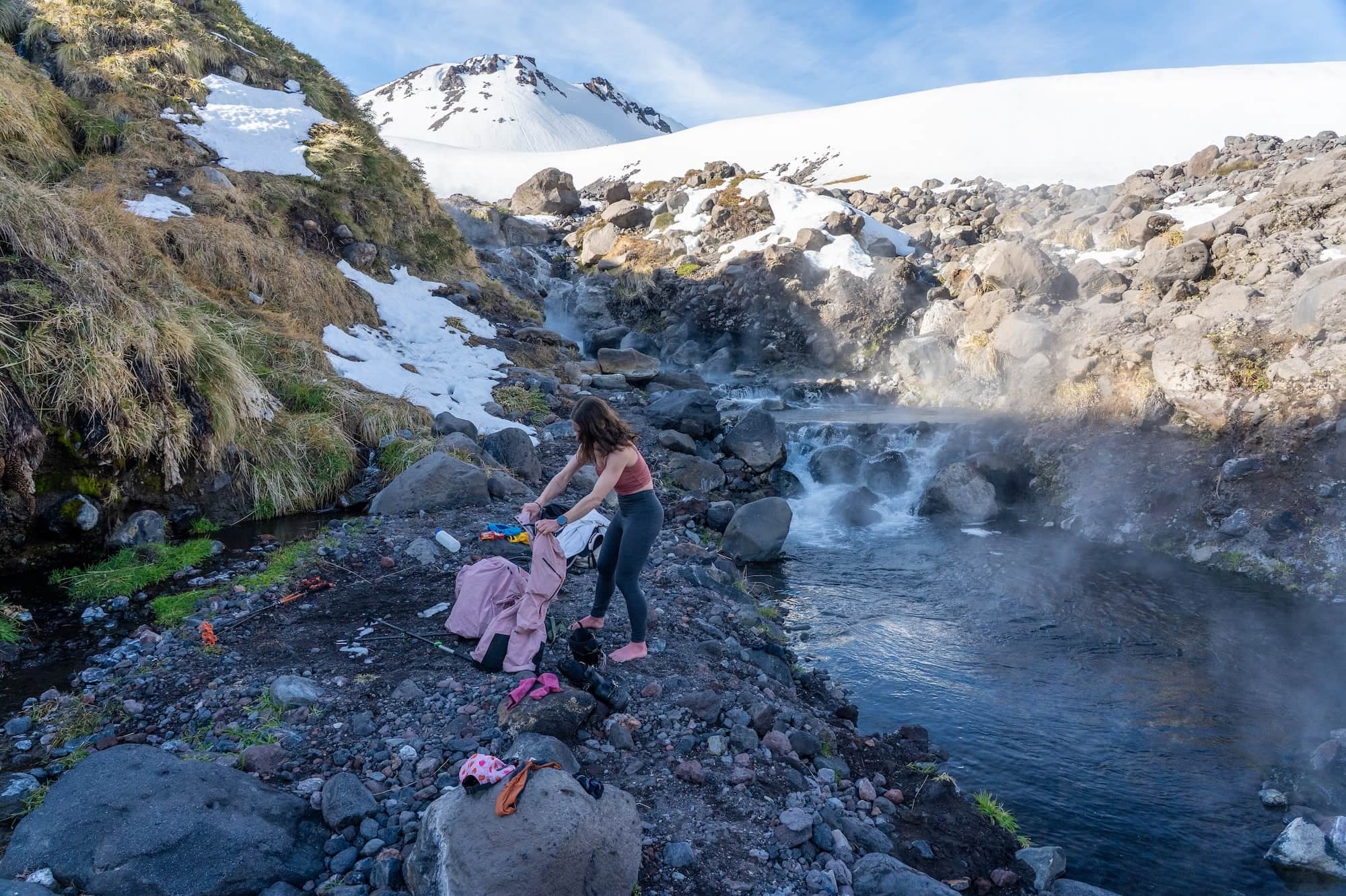 a woman gets undressed by a backcountry hot spring wearing Aspect Base Layer Bottoms and a pink Ridge Merino Bralette