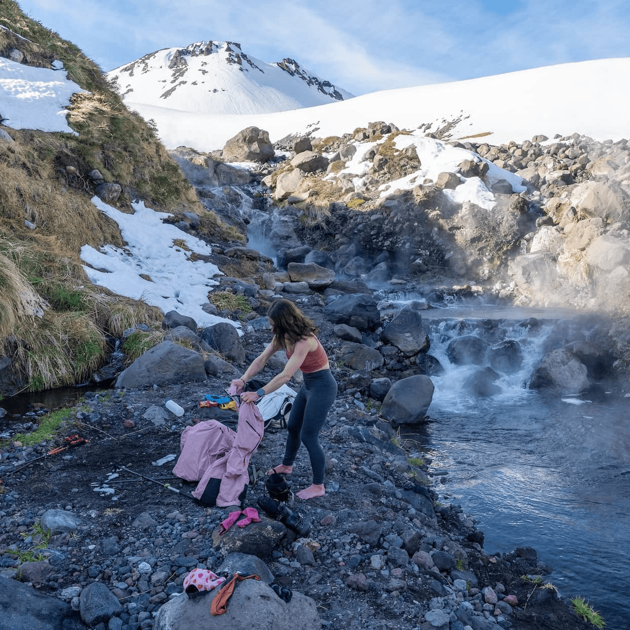 a woman gets undressed by a backcountry hot spring wearing Aspect Base Layer Bottoms and a pink Ridge Merino Bralette