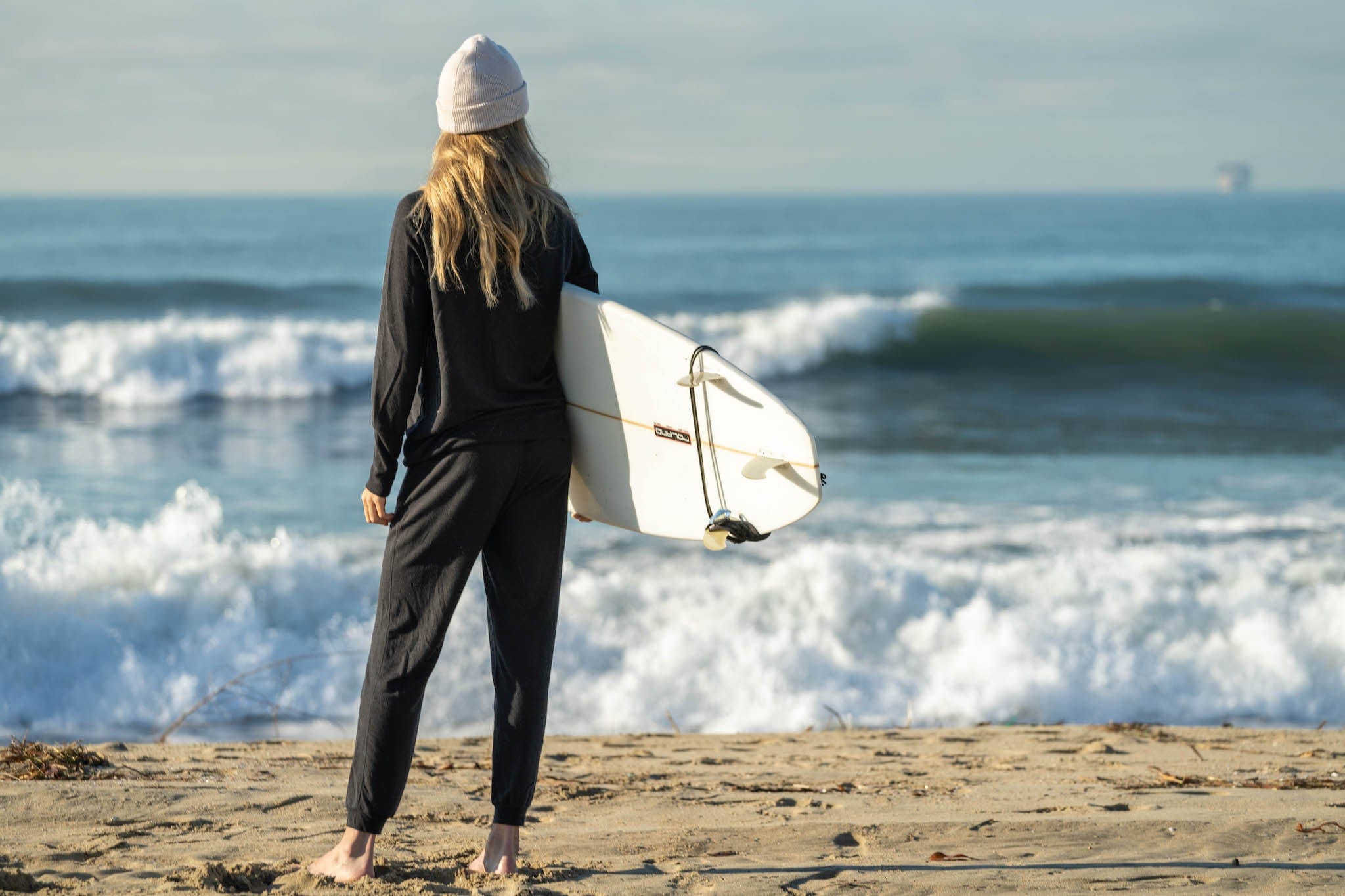Friend of Ridge Merino Shannon Robertson wearing the matching Women's Natural Tencel Joggers and Crew Sweatshirt holding a surfboard in front of the ocean in SoCal