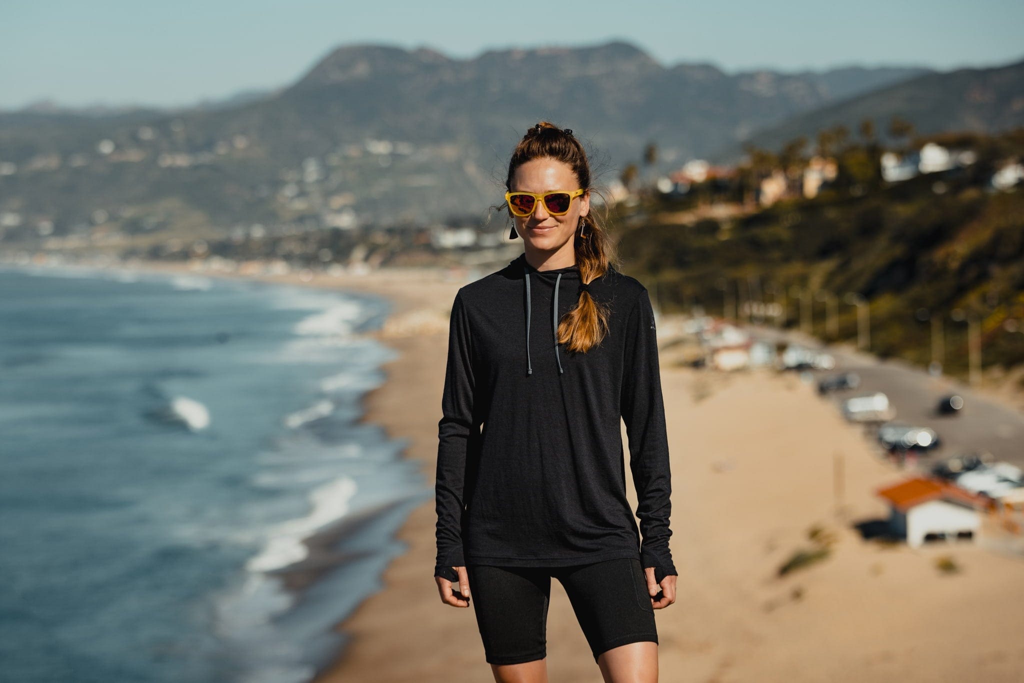 a woman wearing a black Ridge Merino Sun Hoodie and Ridge Hilltop Shorts at the beach on a spring day