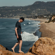 a man wearing a Journey V-Neck Tee walking on rocks above a beach in Malibu