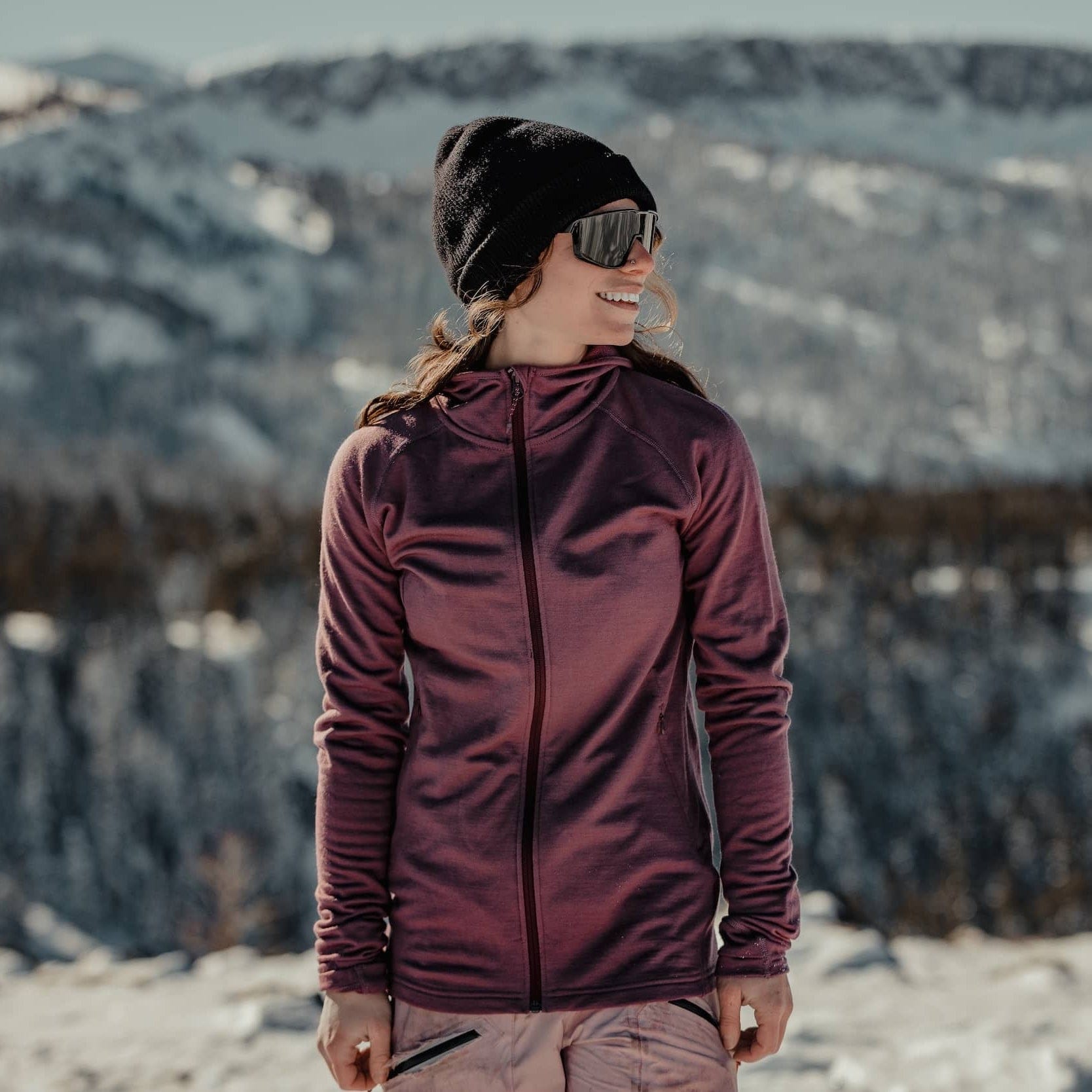 a woman wearing a Hyde Hoodie and Ridge Beanie smiling in a snowy mountain landscape