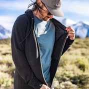 a man unzipping a black Traveler Hoodie over a blue t-shirt in the mountains