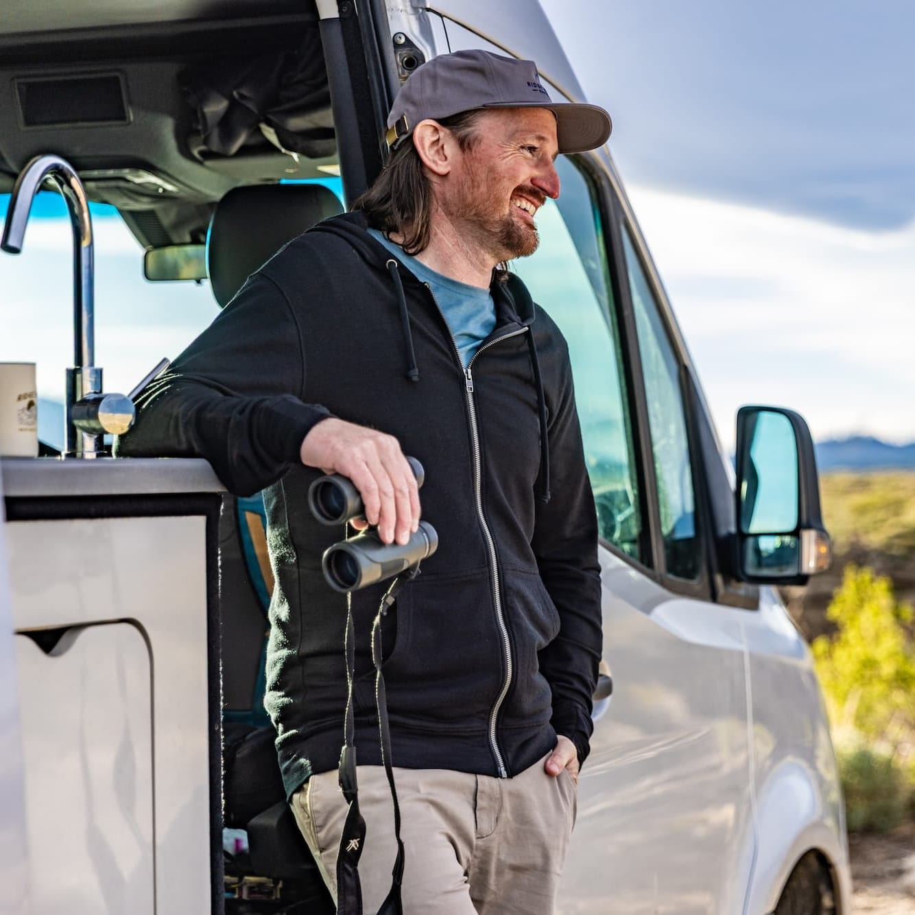 a man stands in his van with binoculars wearing a Traveler Hoodie and Ridge Merino cap