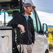 a man stands in his van with binoculars wearing a Traveler Hoodie and Ridge Merino cap