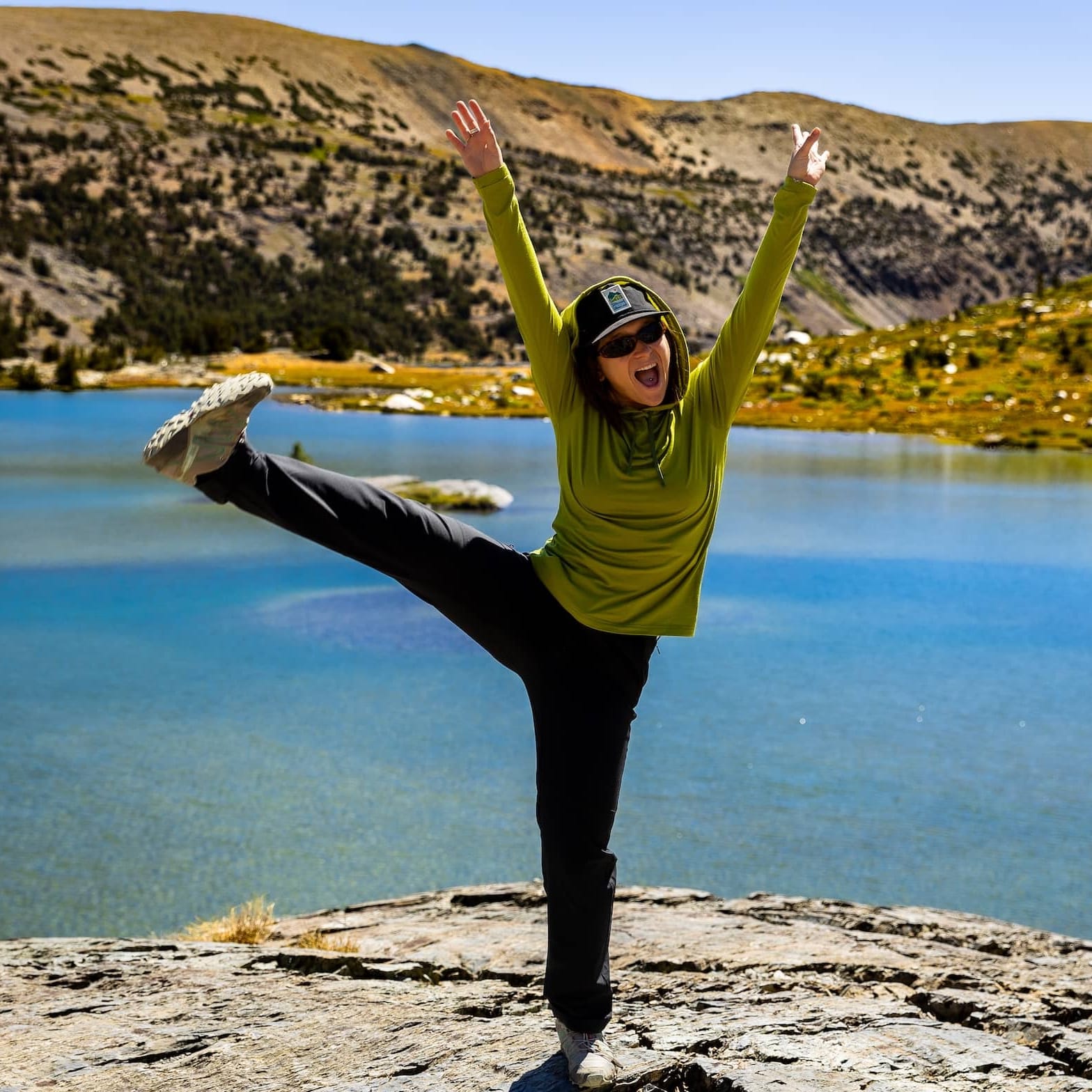 a hiker smiles kicking her leg up wearing a green Merino Wool Sun Hoodie
