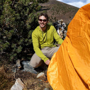 a man smiles setting up a tent wearing a green Solstice Merino Sun Hoodie