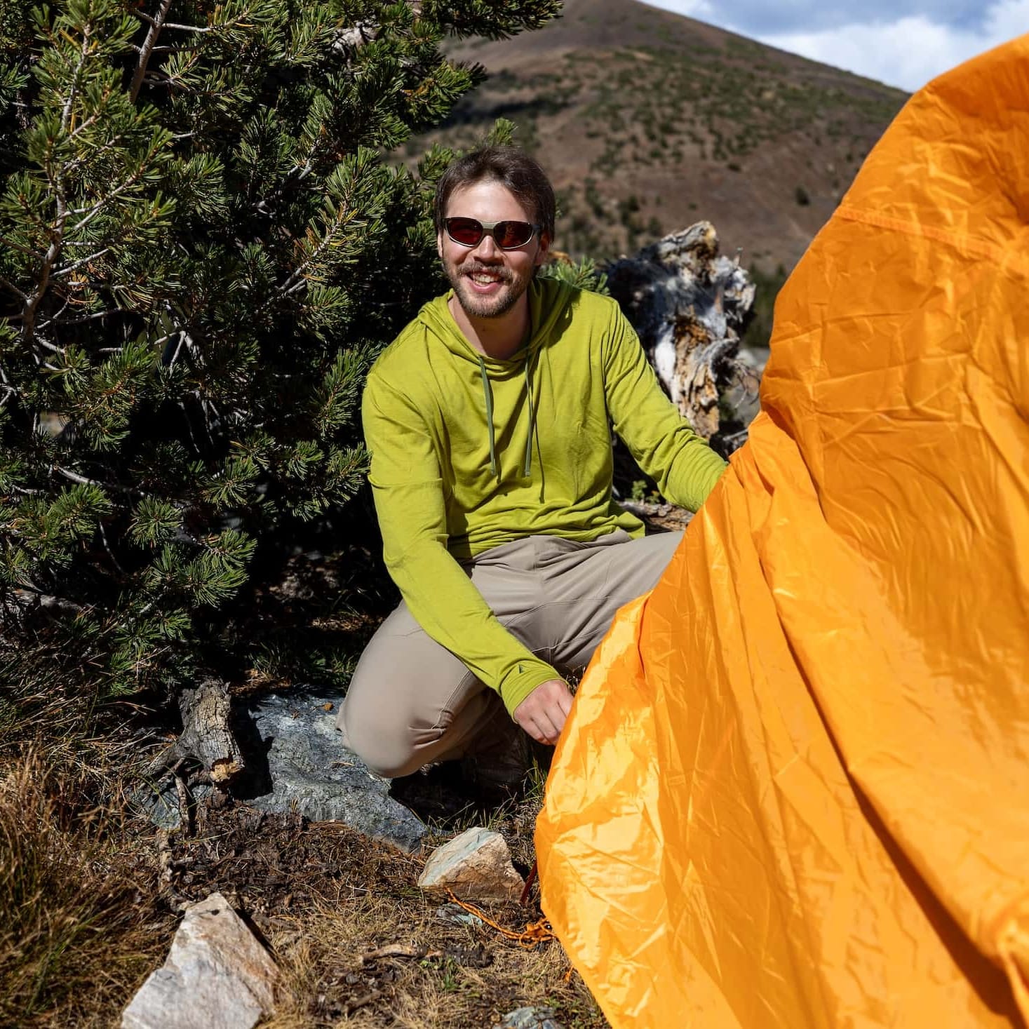 a man smiles setting up a tent wearing a green Solstice Merino Sun Hoodie