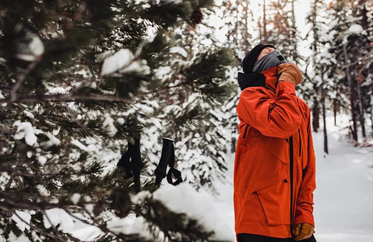 a person zips up their orange ski coat over a Ridge balaclava on a snowy day