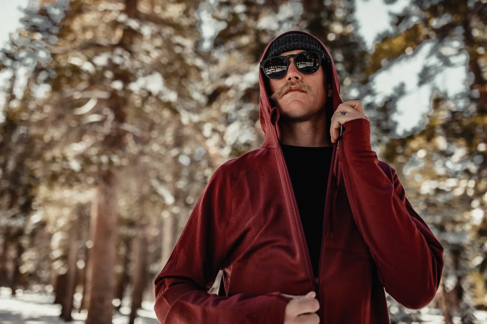 a man stands in a snowy forest wearing a Ridge Merino Hyde midlayer and sunglasses