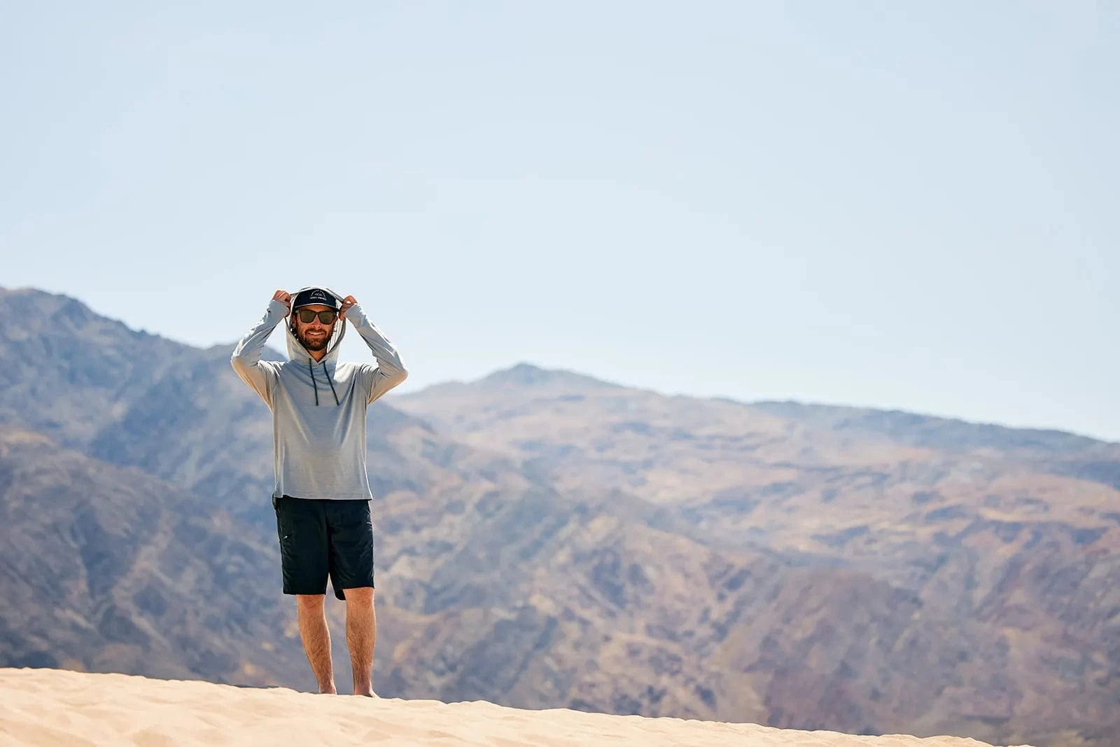 a man stands on a sand dune in Death Valley wearing the Ridge Merino Solstice Sun Hoodie