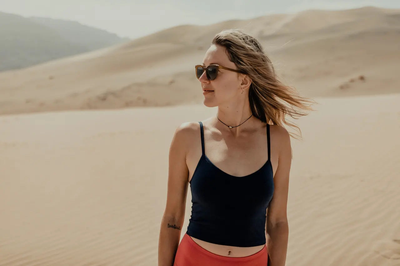 a woman smiles wearing sunglasses and the Ridge Merino bralette on a sand dune